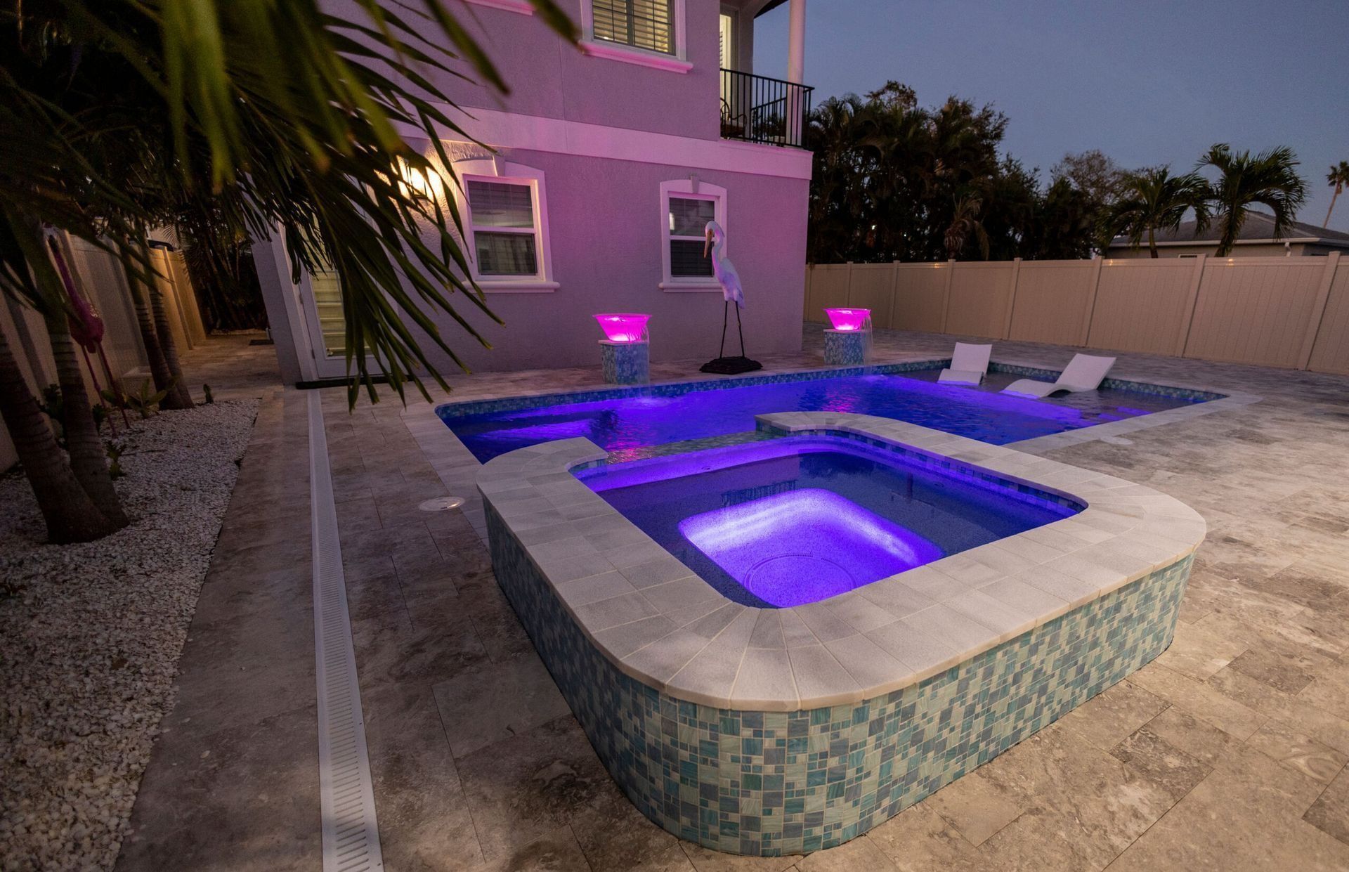Backyard pool at dusk, with purple lighting. Hot tub and lounge chairs are illuminated.
