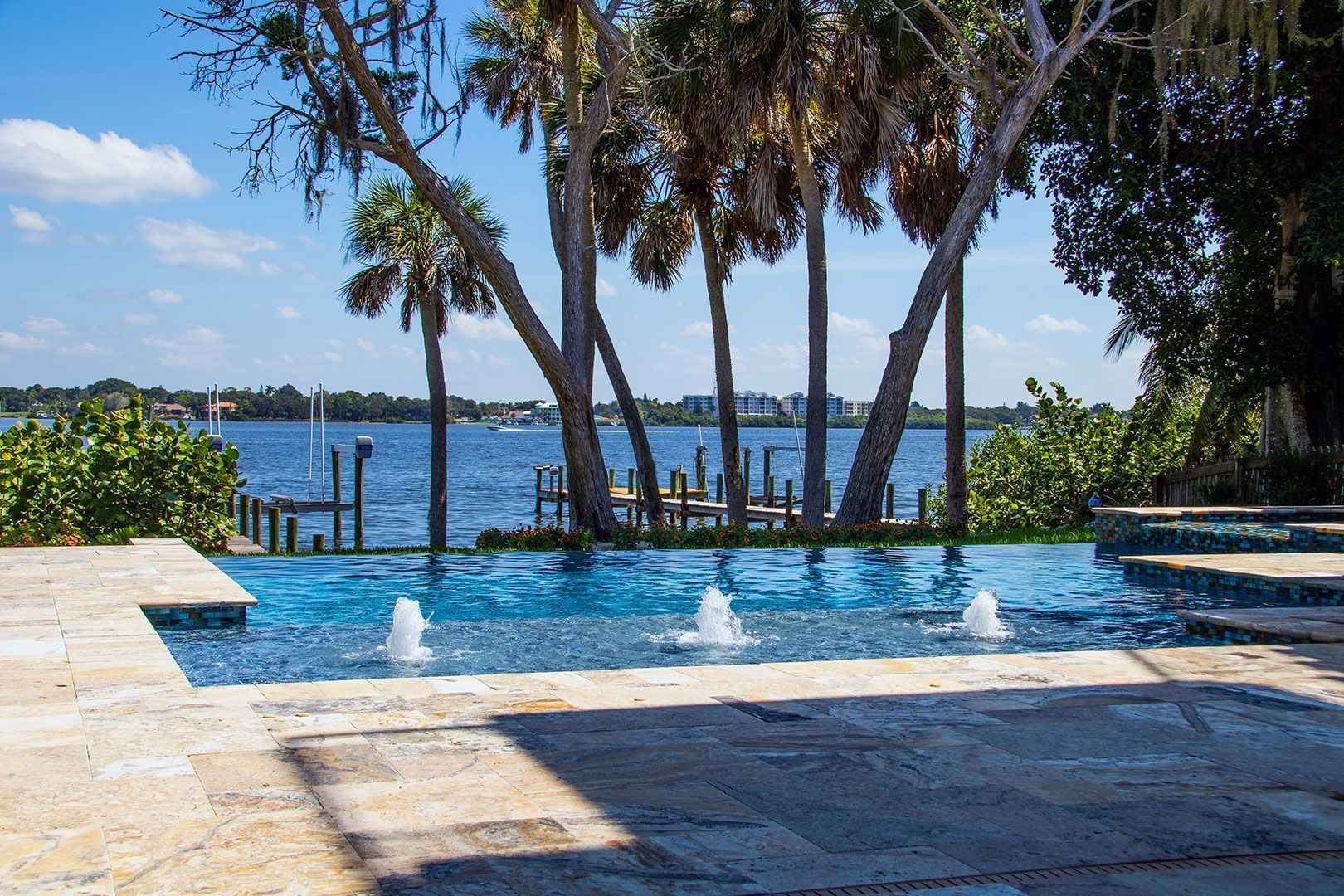 Pool with water fountains overlooking a lake with palm trees and a dock.