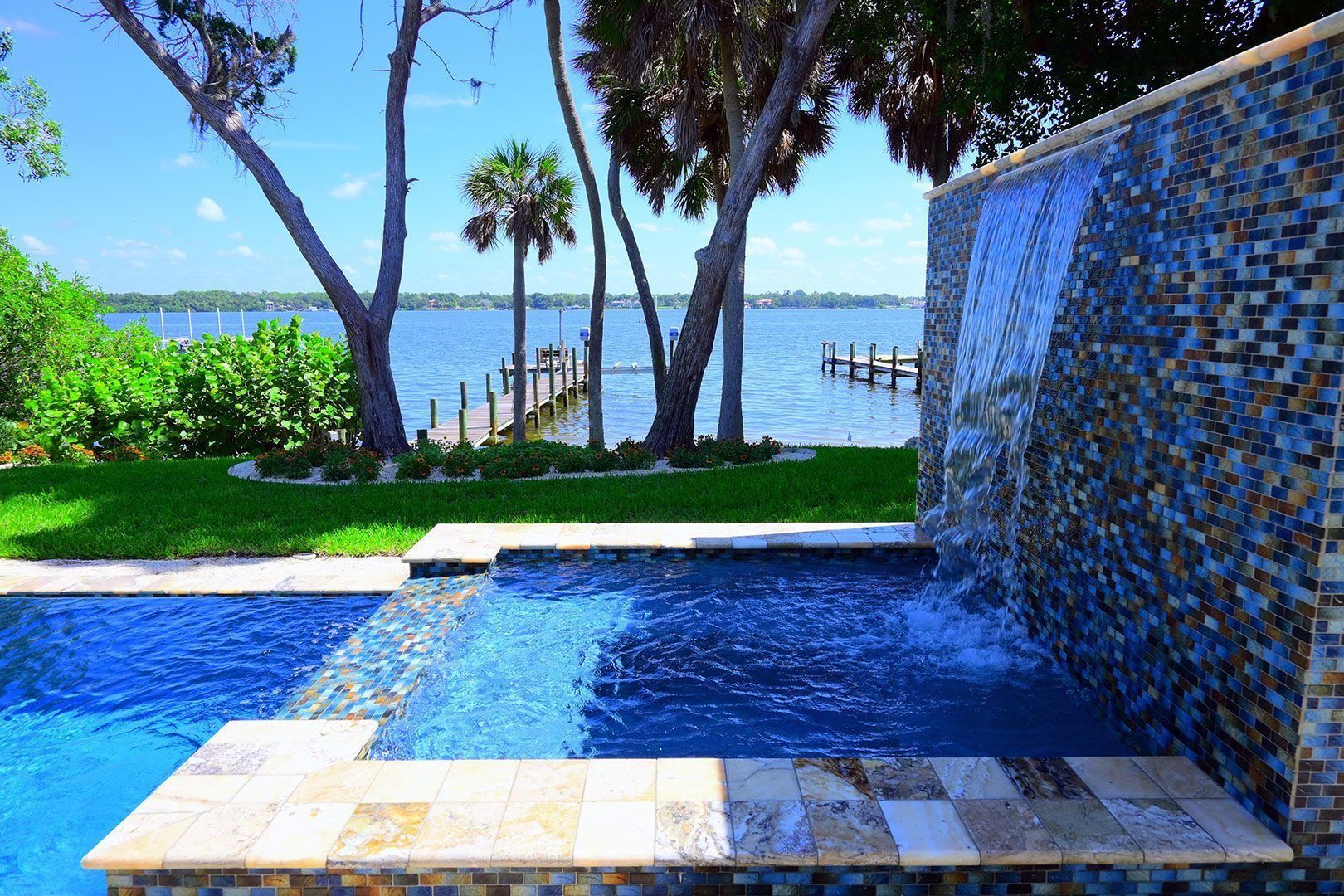 Pool with waterfall feature overlooking a lake, blue water and mosaic tile.