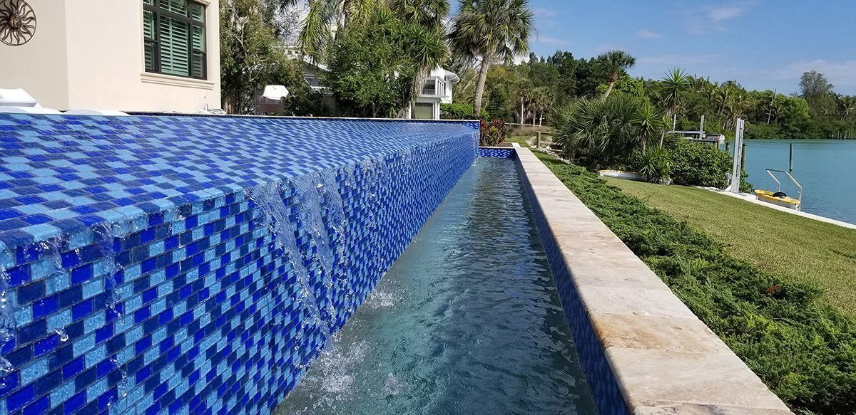Blue tiled infinity pool with water cascading into a trough, overlooking a body of water and greenery.