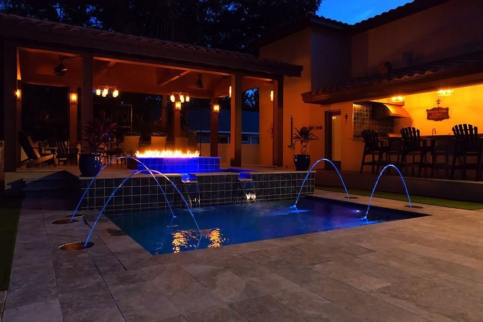 Nighttime outdoor pool with fountain jets, fire pit, and covered bar area illuminated with blue and warm lights.
