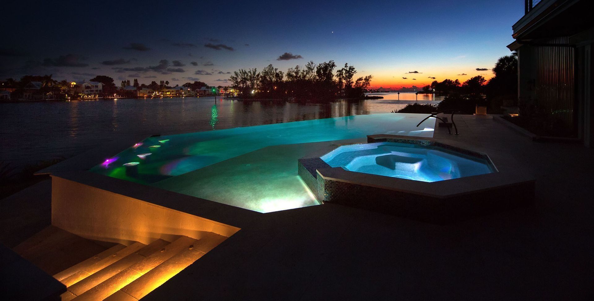 Night view of an infinity pool and hot tub illuminated with blue and green lights, overlooking a body of water at dusk.