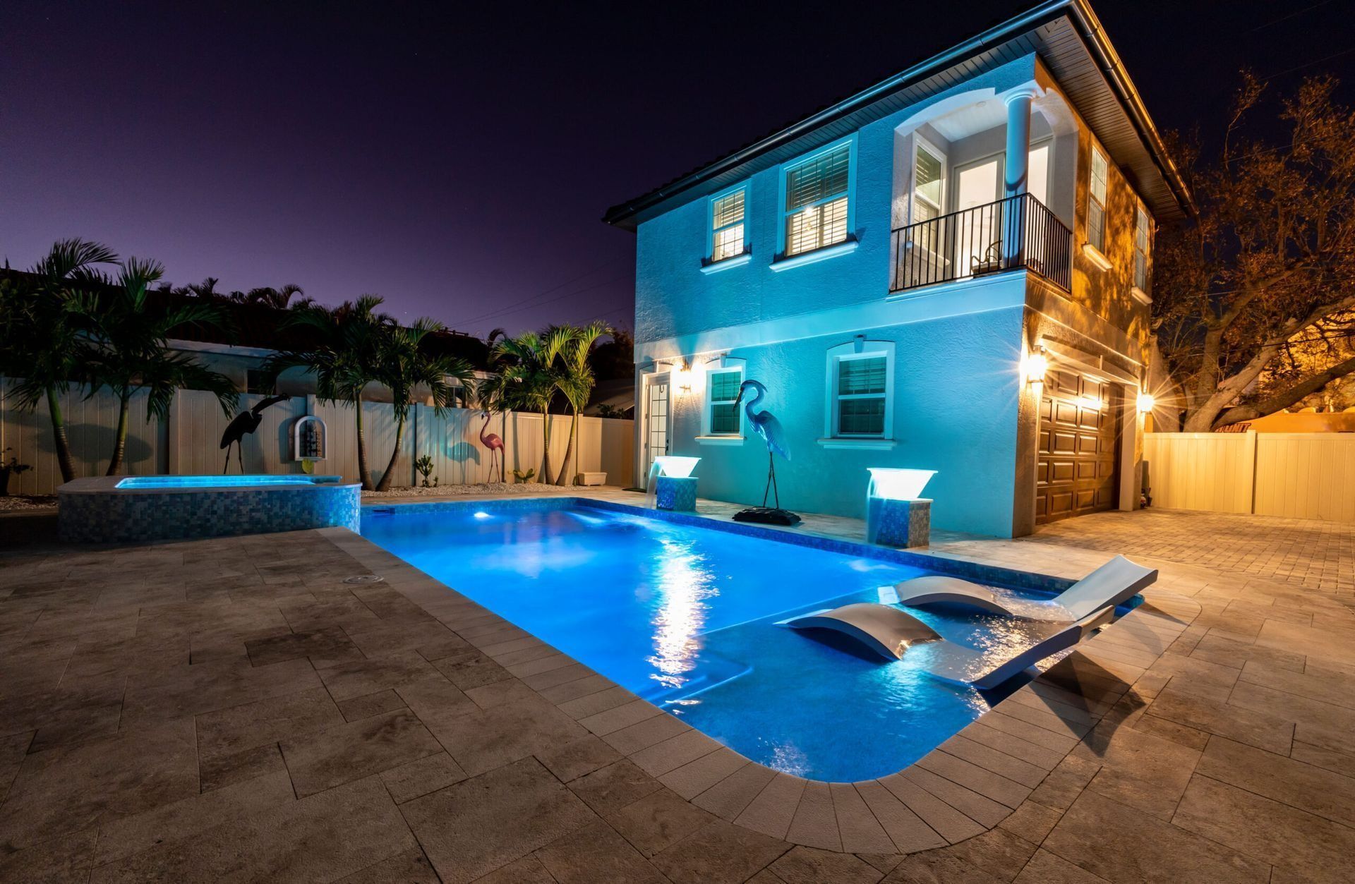 Blue-lit pool at night with two-story house. Loungers in pool. Palm trees and a hot tub.