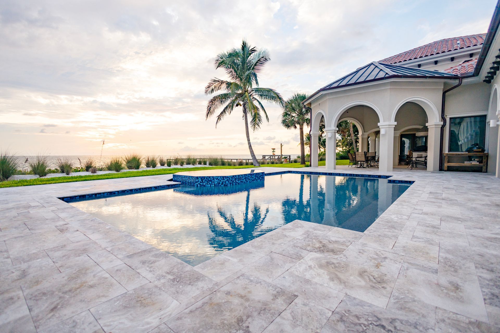 Luxury home with pool overlooking ocean at sunset. Palm trees, blue water, and beige patio.