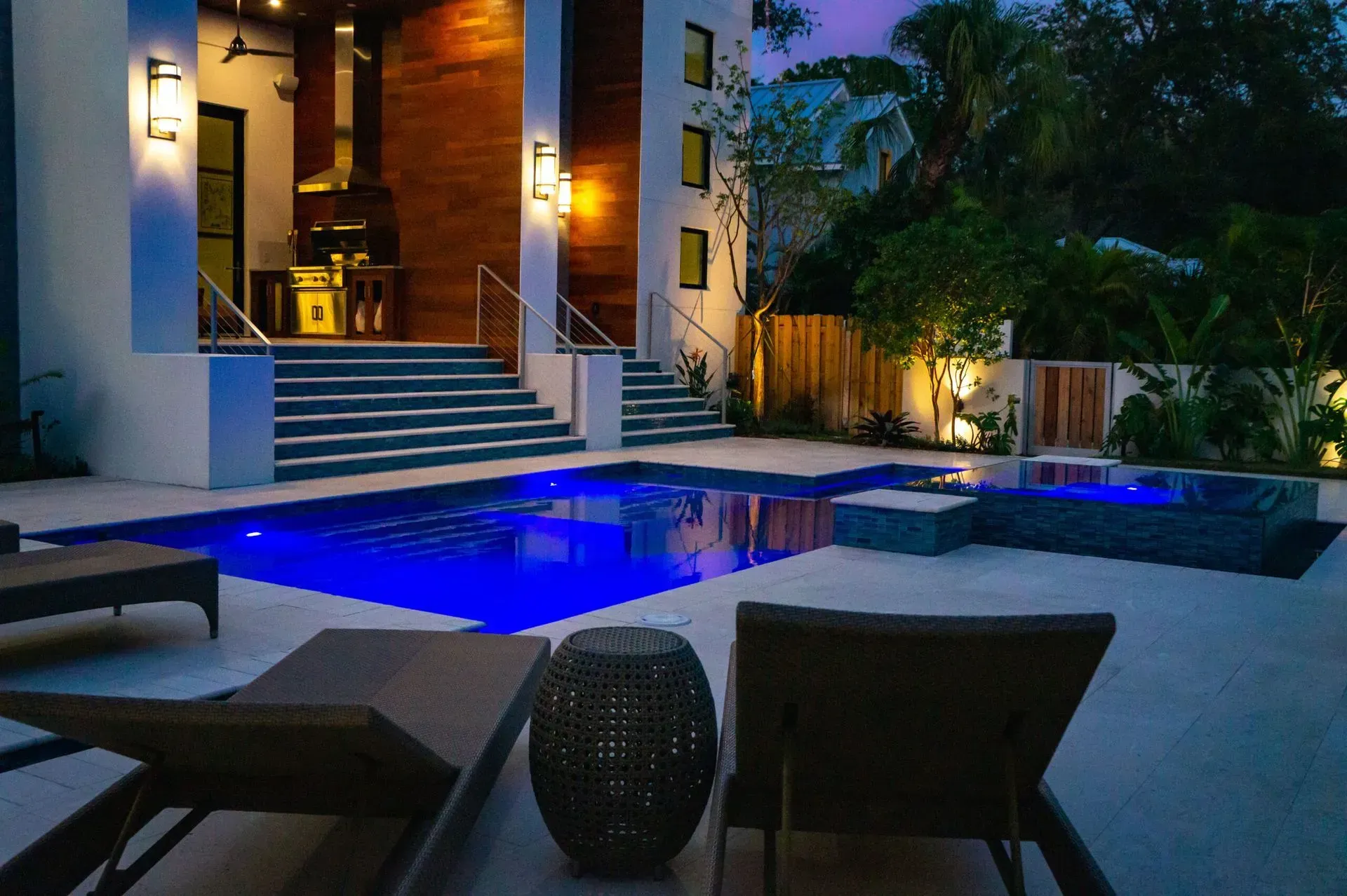 Nighttime view of a luxurious backyard with a lit pool, lounge chairs, and a modern home.