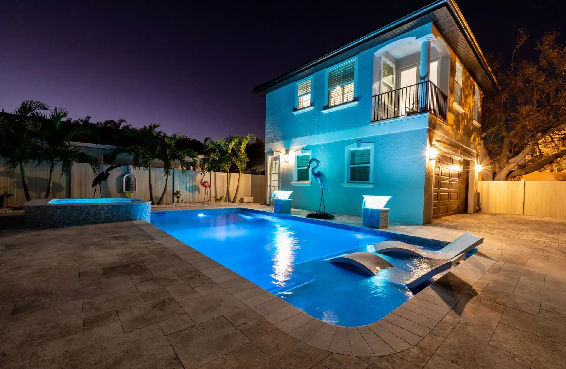 A two-story blue house at night, with a lit pool and patio, and palm trees.