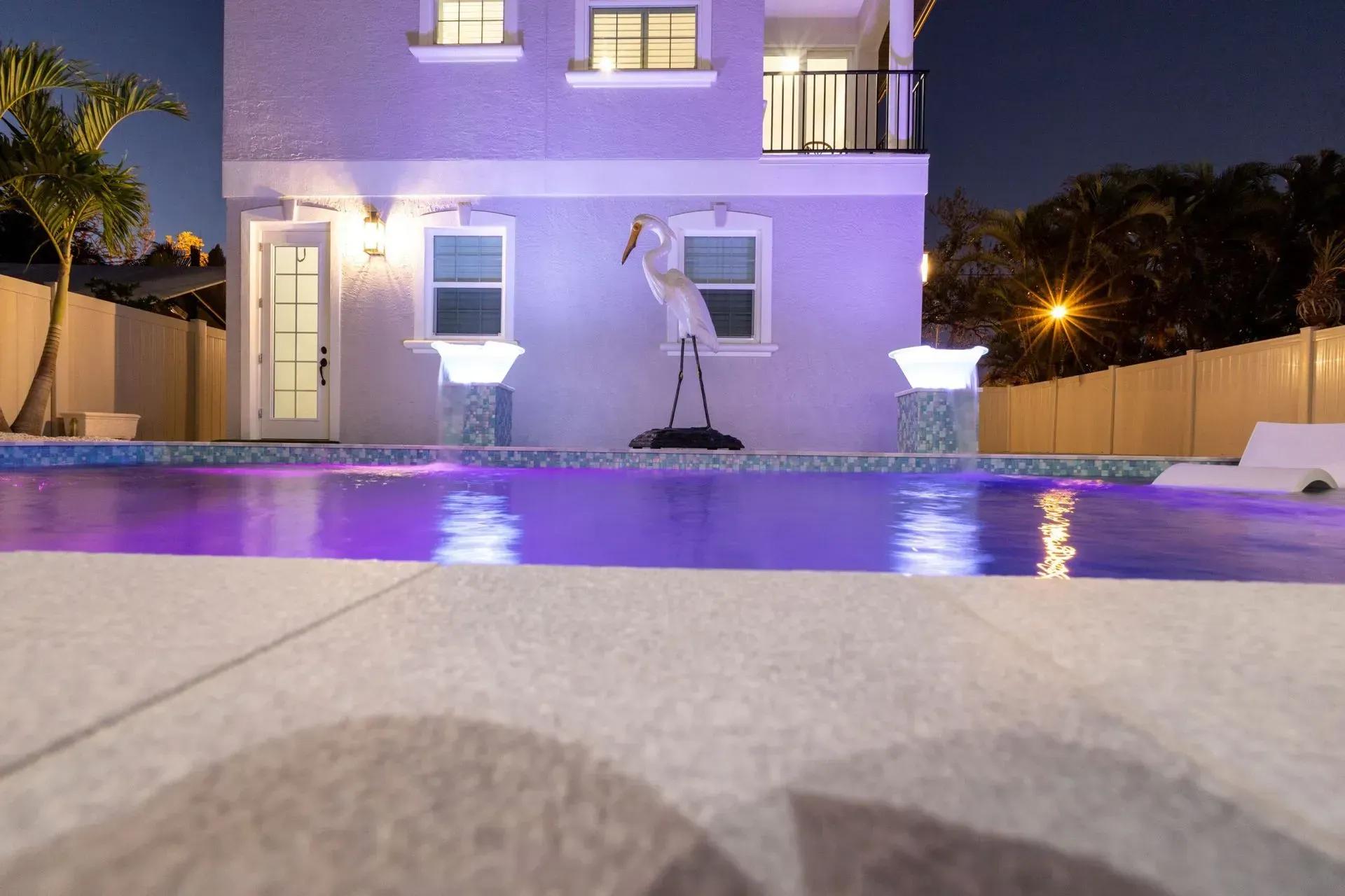 Night view of a house with a pool illuminated in purple; statue of a heron stands in the center.