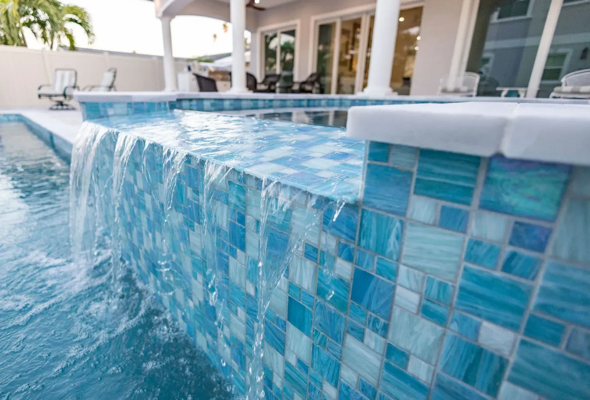 Close-up of a pool with a tiled waterfall. The tiles are blue and water cascades down. A house is in the background.