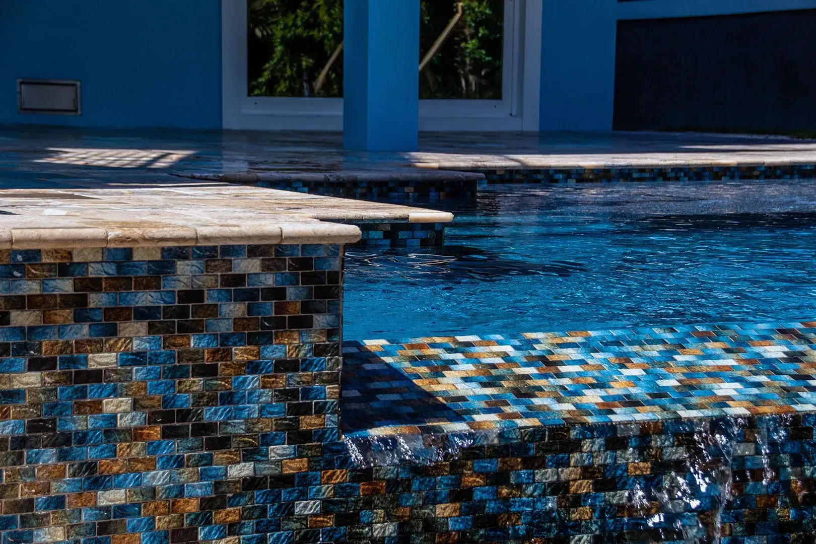 Pool with blue water, blue and brown tile, and a blue building in the background.