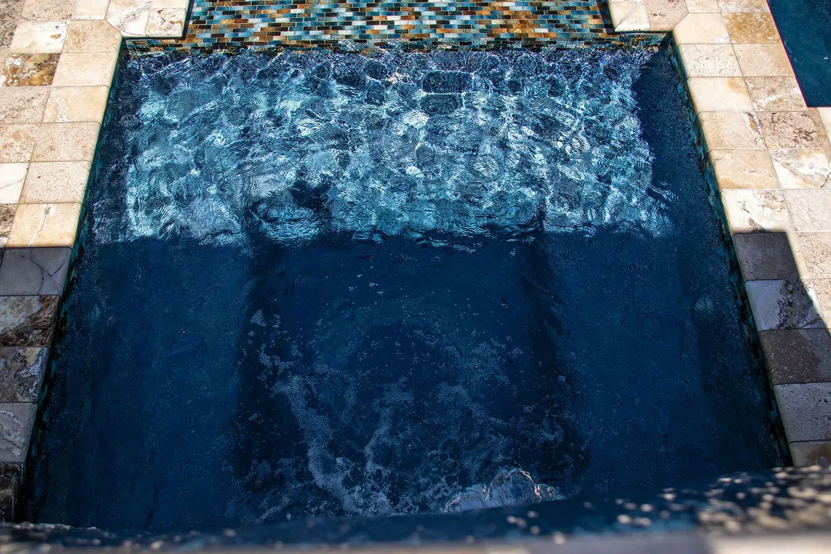 Blue tiled pool with water cascading into it, surrounded by stone tiles.