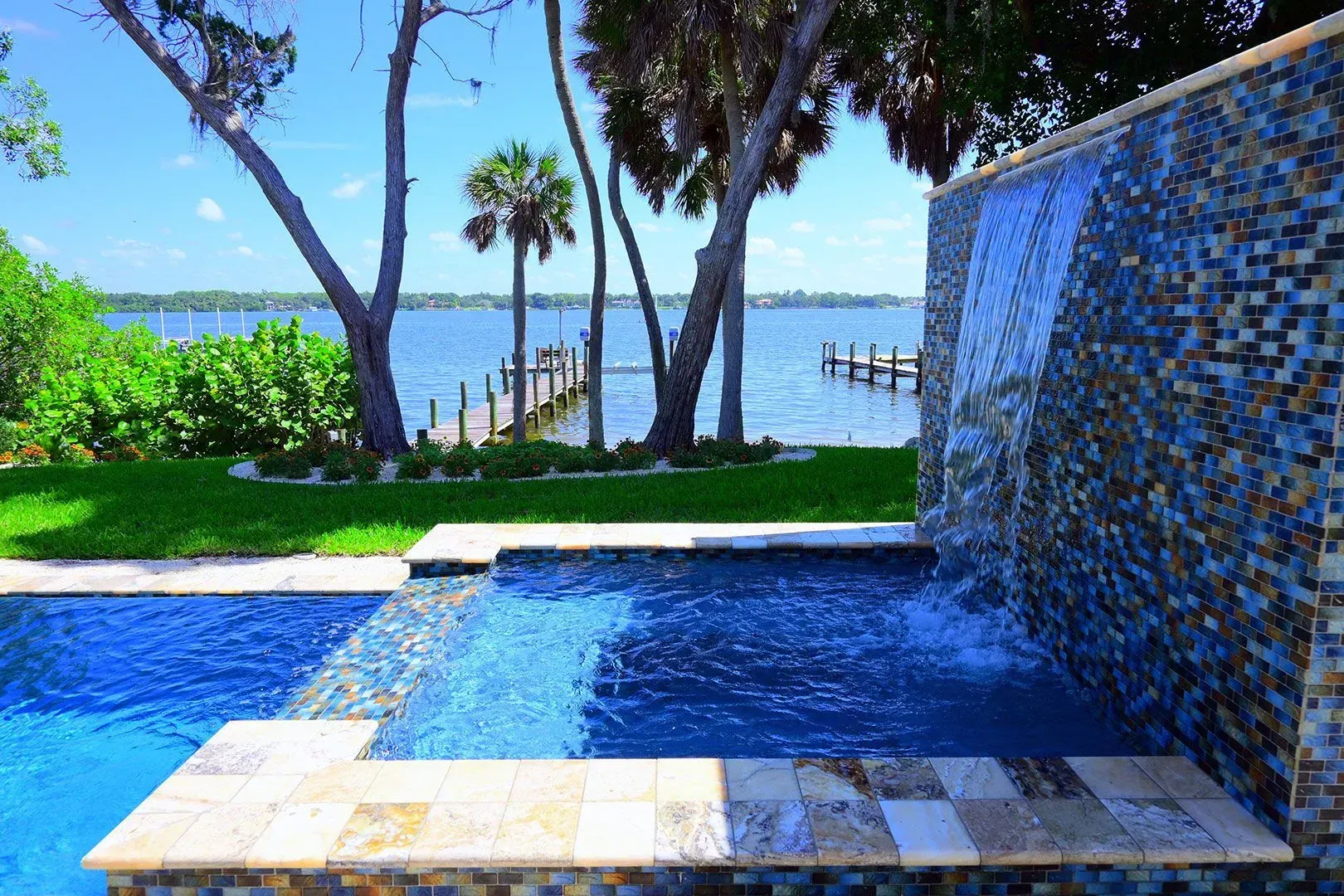 Pool with waterfall feature overlooking a body of water with a dock and trees.