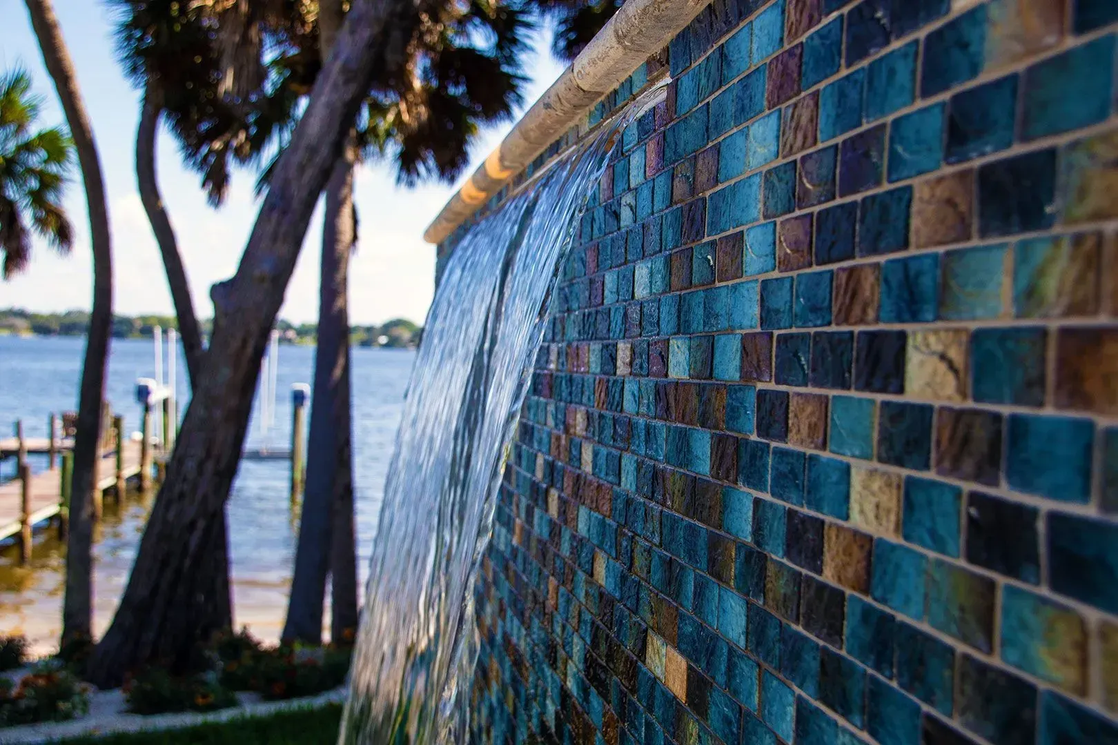 Water cascading down a blue and brown tiled wall with palm trees and a waterfront in the background.
