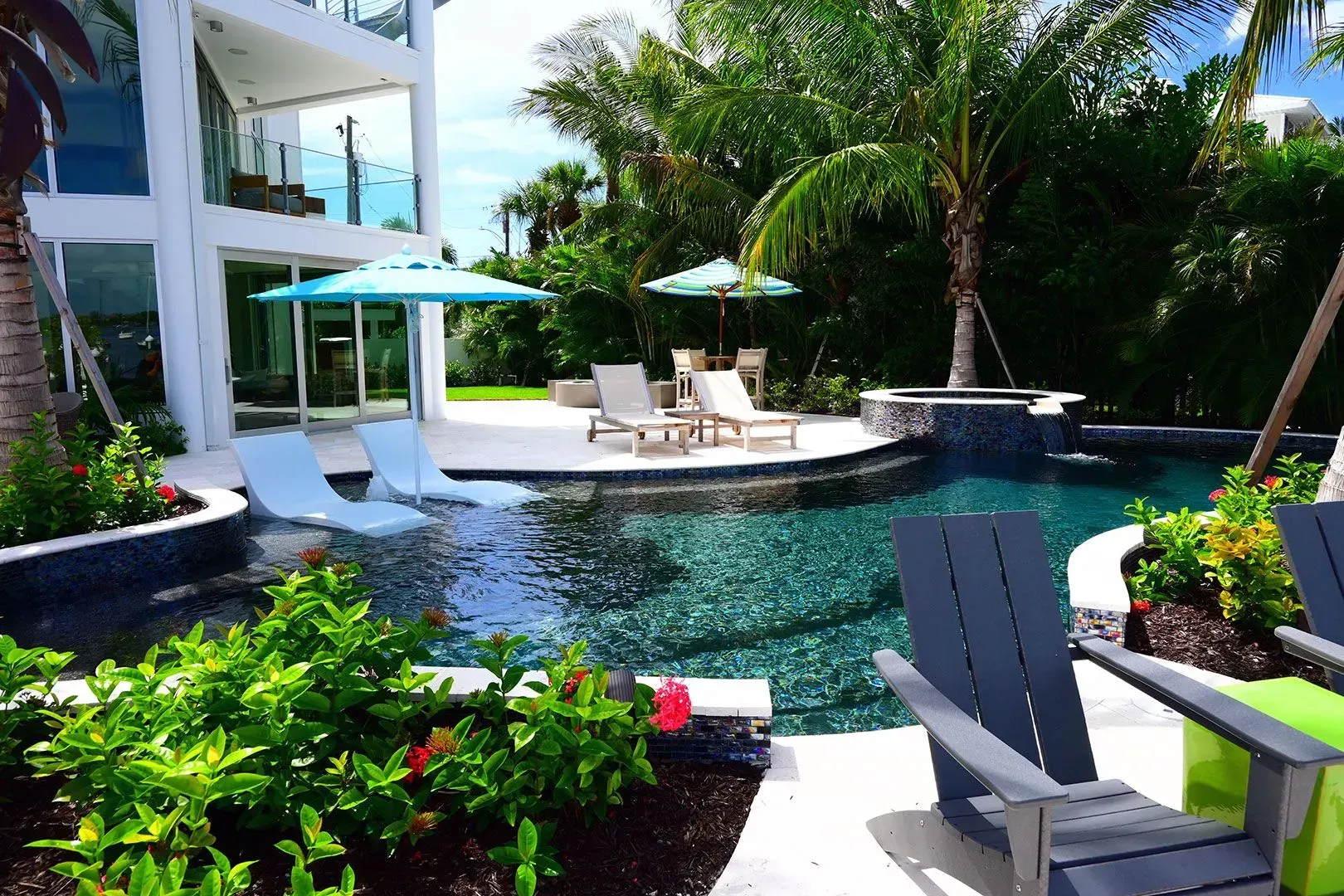 Luxury backyard with pool, lounge chairs, umbrellas, and modern white house. Tropical plants and blue water.