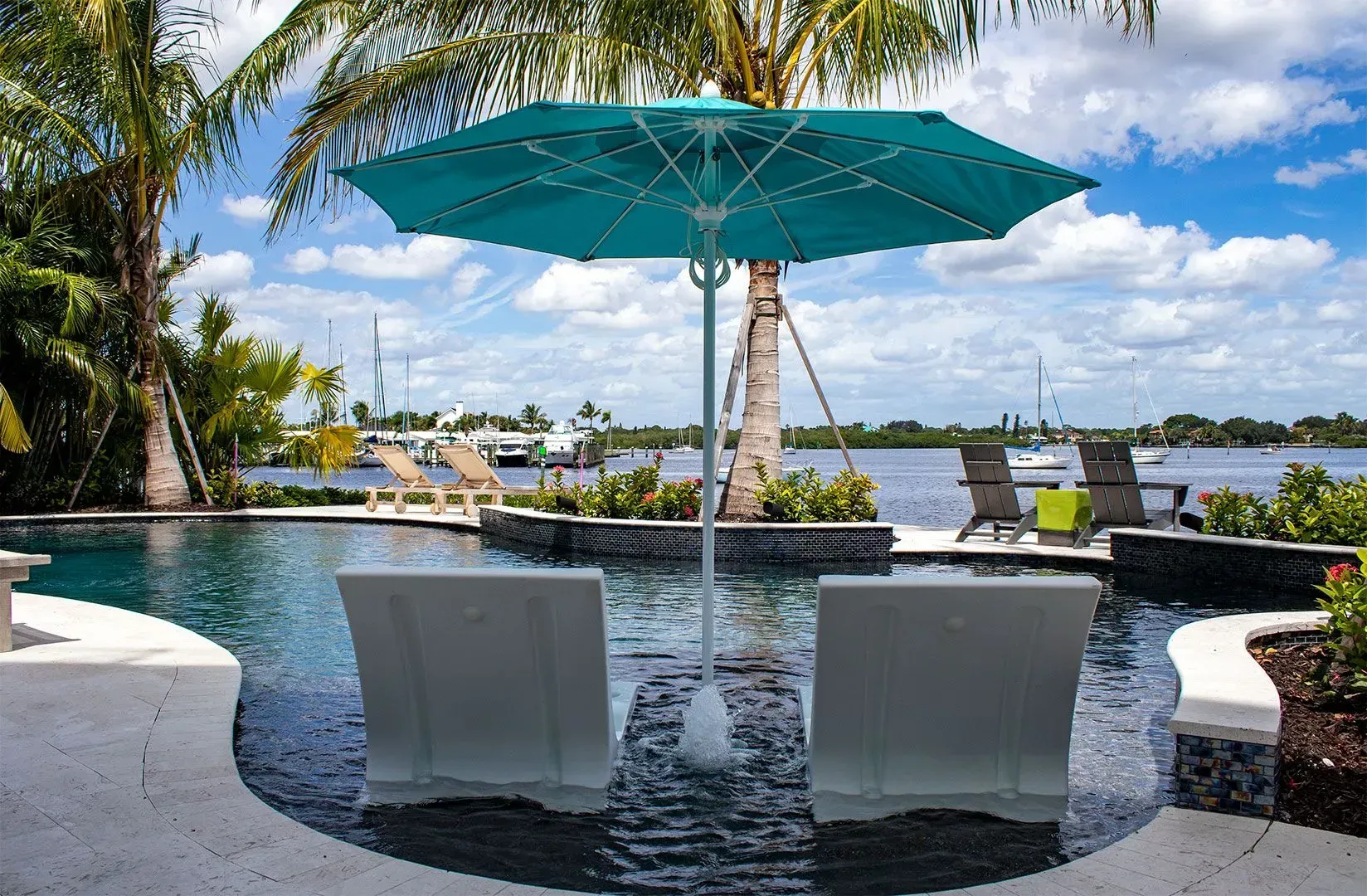 Poolside scene with turquoise umbrella over two submerged white chairs, overlooking water and boats.
