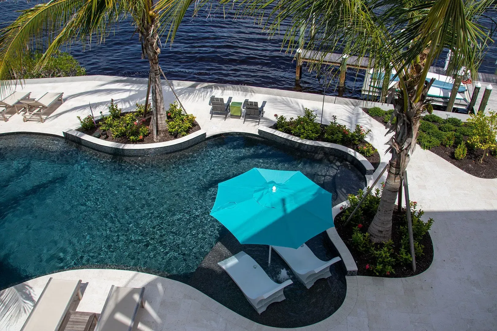Pool with turquoise umbrella and lounge chairs, near a body of water with docks.