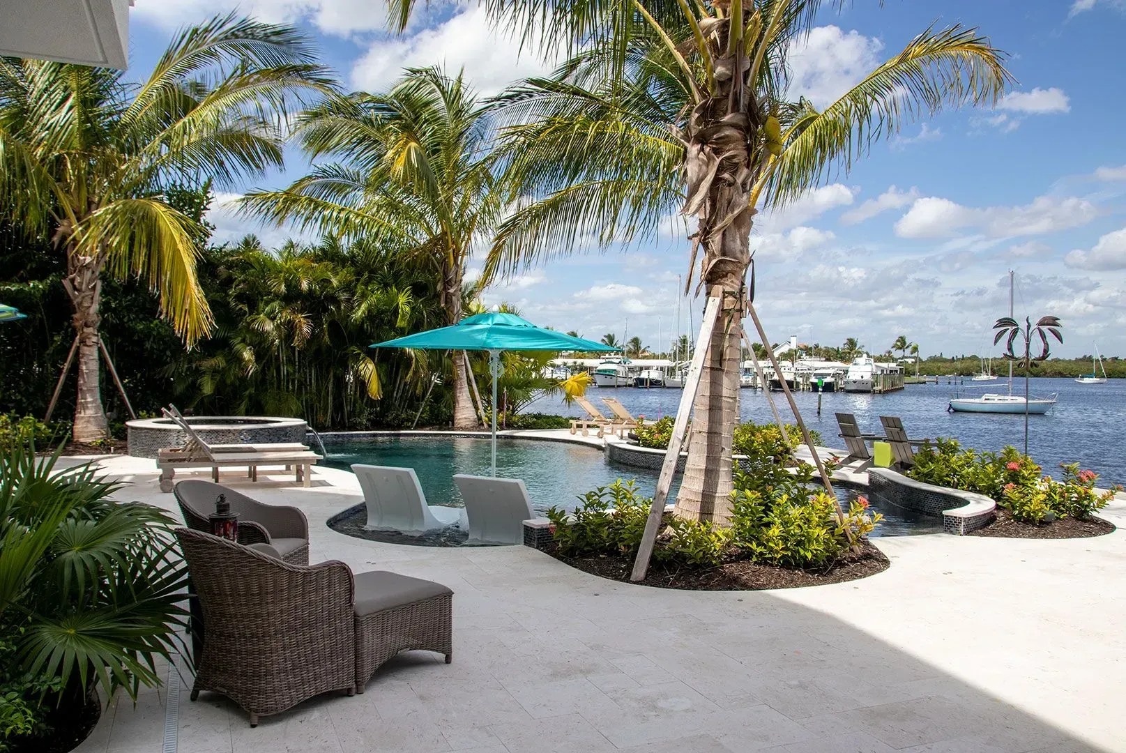 Patio with pool and water view, palm trees, lounge chairs, sunny day.