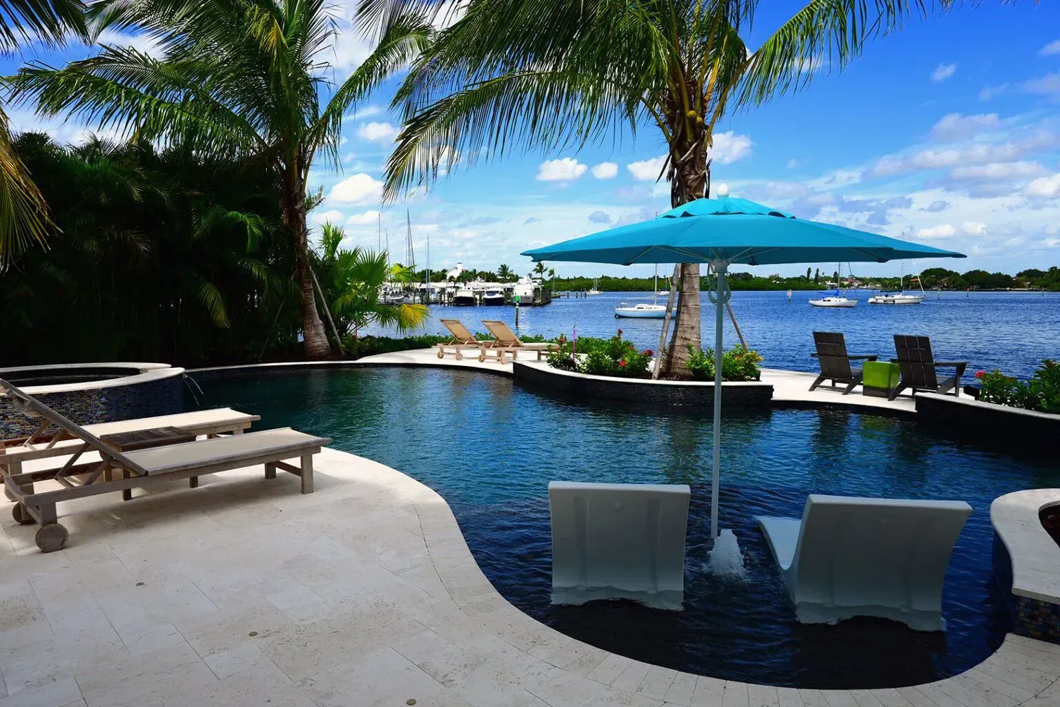 Poolside lounge area with blue water, turquoise umbrella, palm trees, and water view.