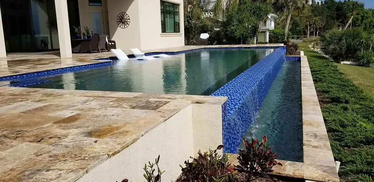 A pool with blue tile and water feature, next to a home and green plants.