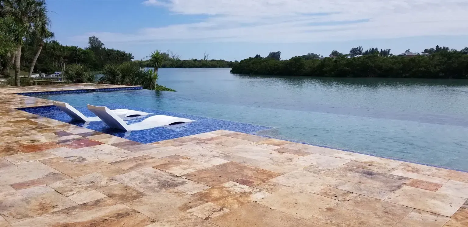 Pool with blue tiles and white lounge chairs next to a blue waterway lined with trees.