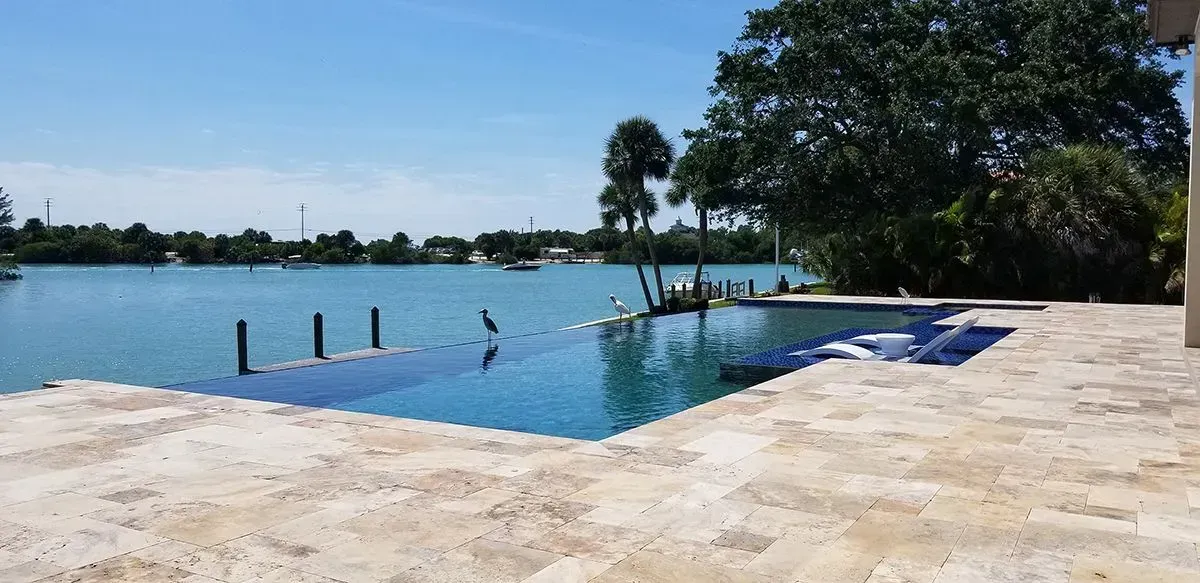A pool with blue tile overlooking a body of water. Trees and a clear blue sky in the background.