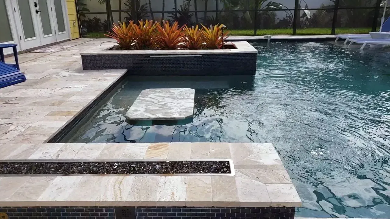 Pool with submerged bar, seating, and planter with colorful foliage. Beige stone, blue tile, and clear water.