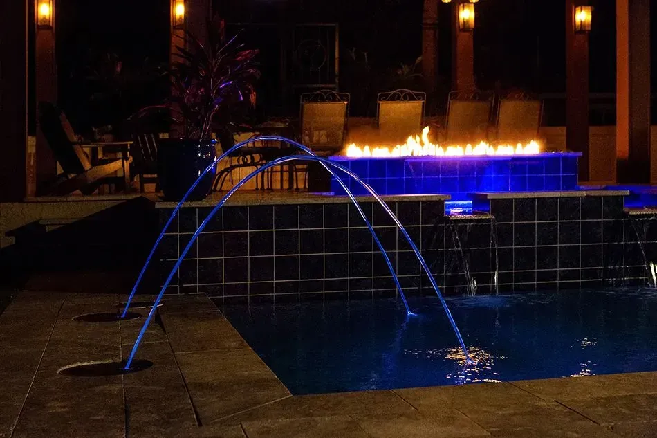 A nighttime pool with blue-lit water fountains and a fire pit in the background.