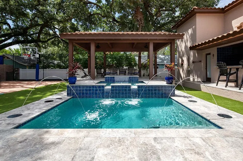 Backyard pool with fountains and a pergola. Blue water, tan patio, lush green grass, and a basketball hoop.