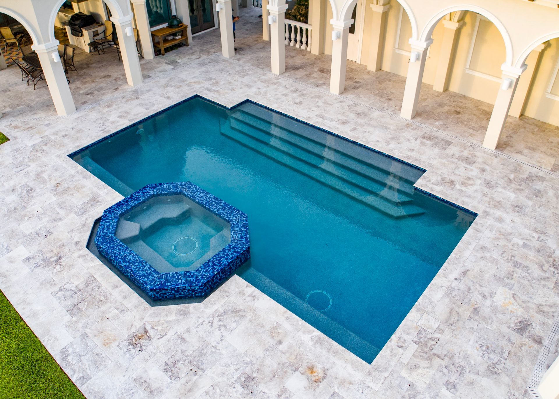 Outdoor pool with steps and attached spa. Blue water, cream-colored patio, white arches.