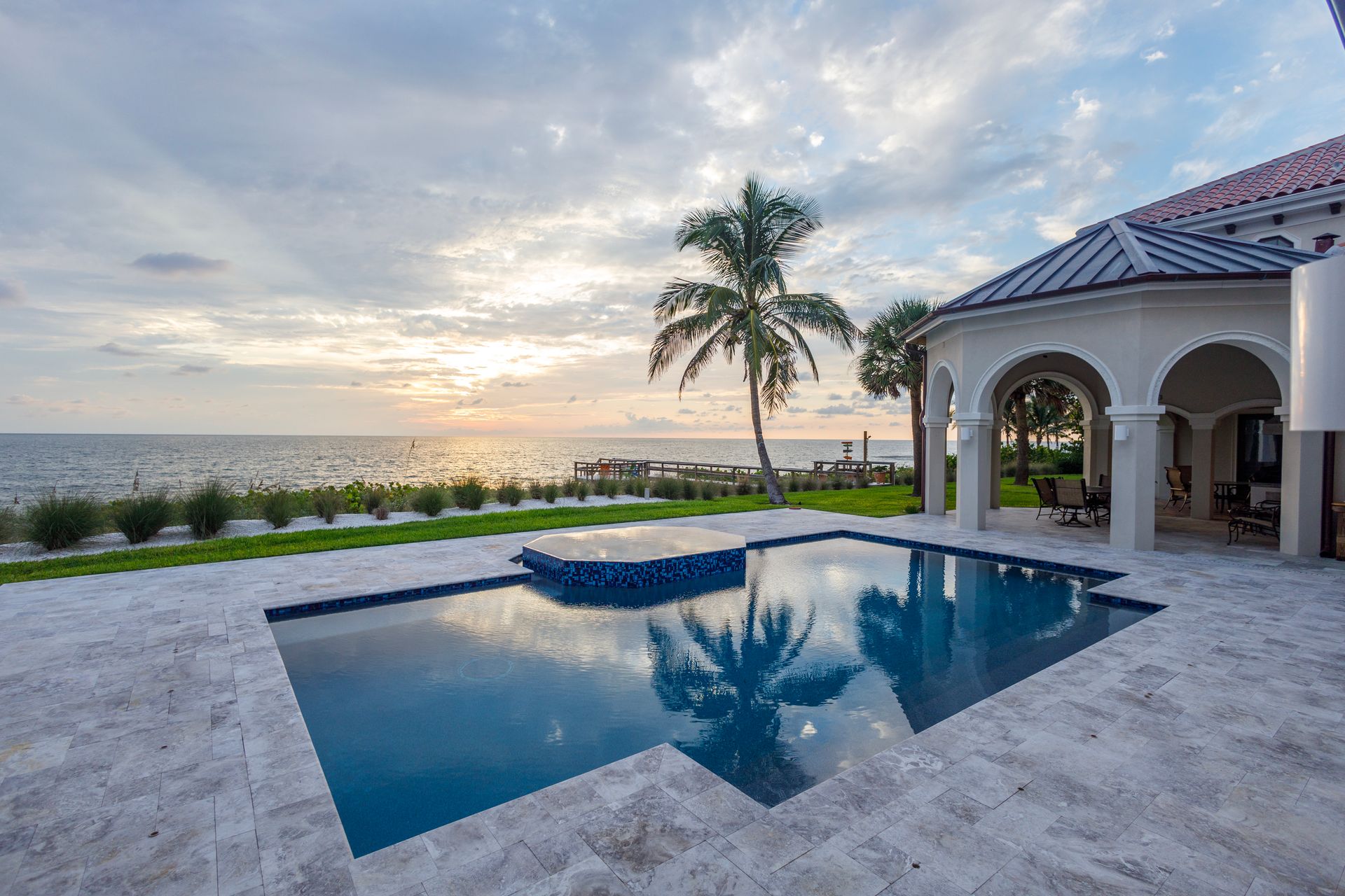 Pool overlooking the ocean at sunset. Palm tree, house with arches, blue water reflecting sky.