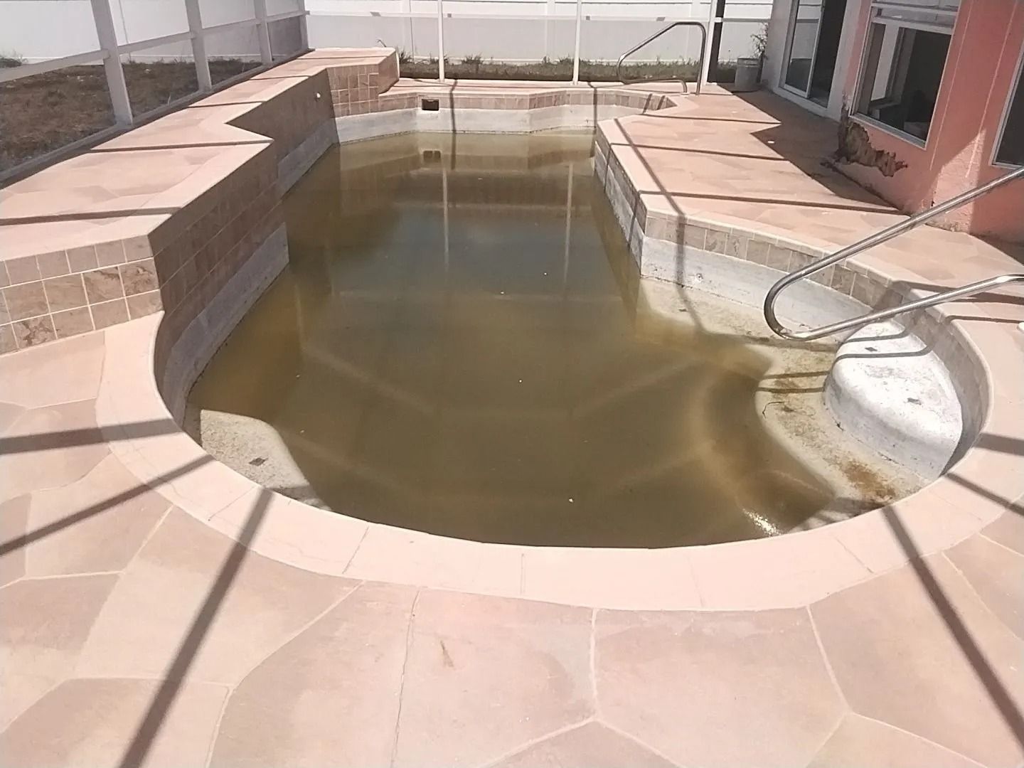 Dirty pool with brown water, surrounded by tan tiles and a pink building.