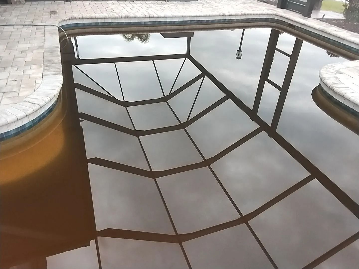 Brown pool water reflecting a screened enclosure and cloudy sky. Brick patio surrounds the pool.