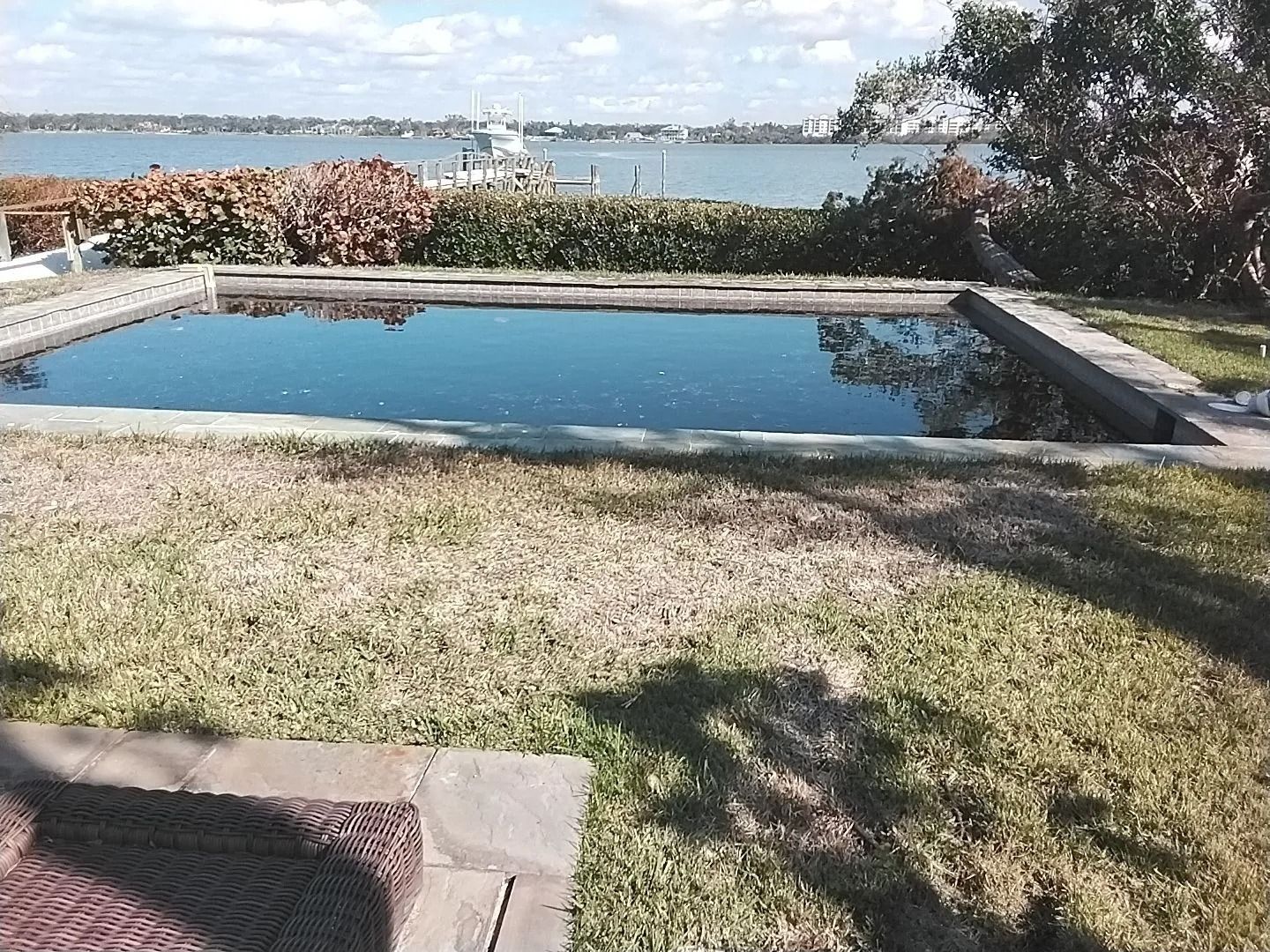 A rectangular, dark-water pool bordered by concrete sits in front of dry grass, with a waterfront view in the distance.