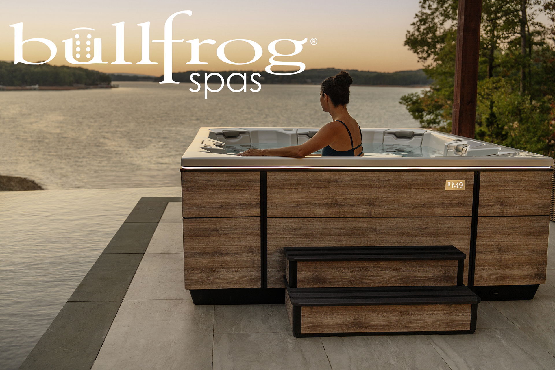 Woman relaxing in a Bullfrog Spas hot tub overlooking a lake at sunset.