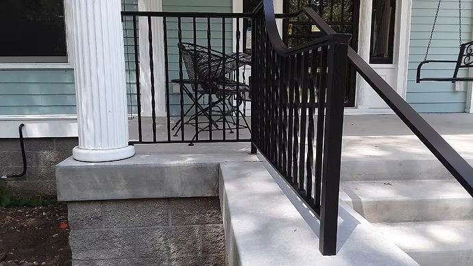 Front porch steps with black railing beside a white column and brick house exterior