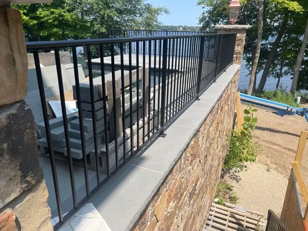 Patio with black railing overlooking a pool and trees beside a stone wall