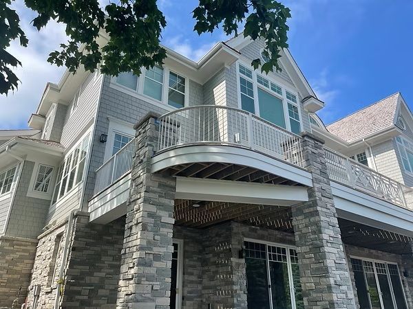 Two-story gray house with stone facade, arched balcony, and white trim under a blue sky