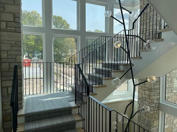 Bright foyer with stairs, black railings, tall windows, and stone walls bathed in sunlight.