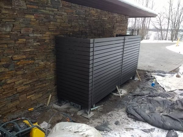 Black slatted storage cabinet beside a stone wall, with snow and plastic sheeting on the ground.