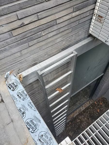 Exterior wall with a mounted metal ladder and vent beside a building window well