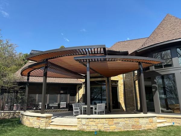 Patio with curved brown pergola, stone wall, and outdoor seating beside a building