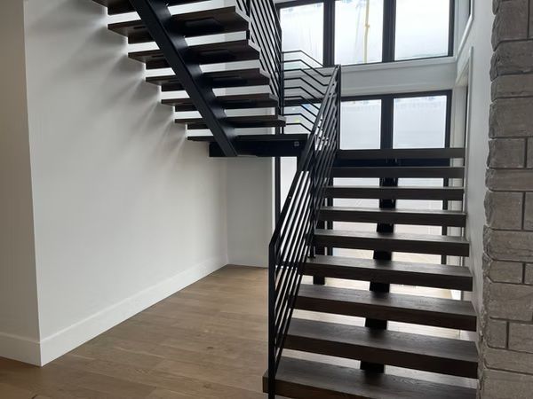 Modern indoor staircase with black metal railings, open risers, and large windows overlooking a wood floor room