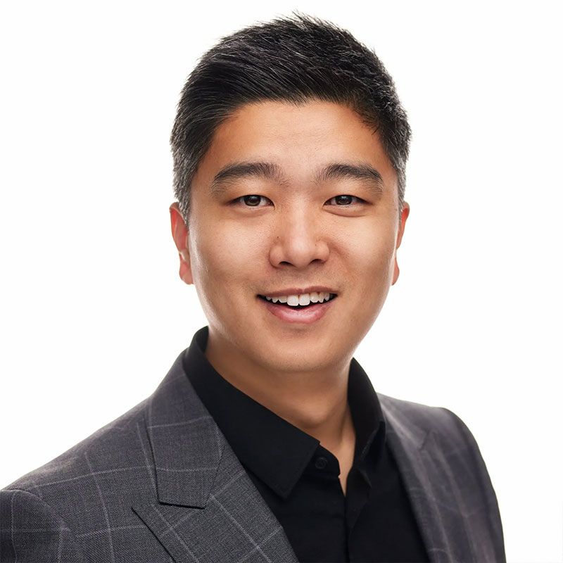 Asian man in gray suit and black shirt smiling at the camera against white background.