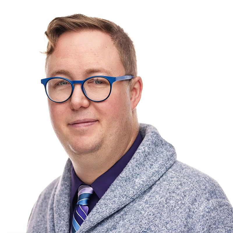Man with glasses and grey cardigan smiles at camera.