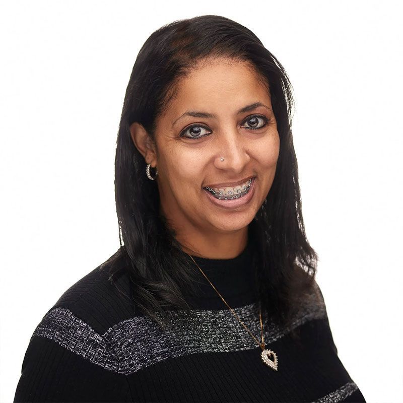 Woman with braces smiles, wearing black sweater with silver stripes, and a heart pendant necklace.