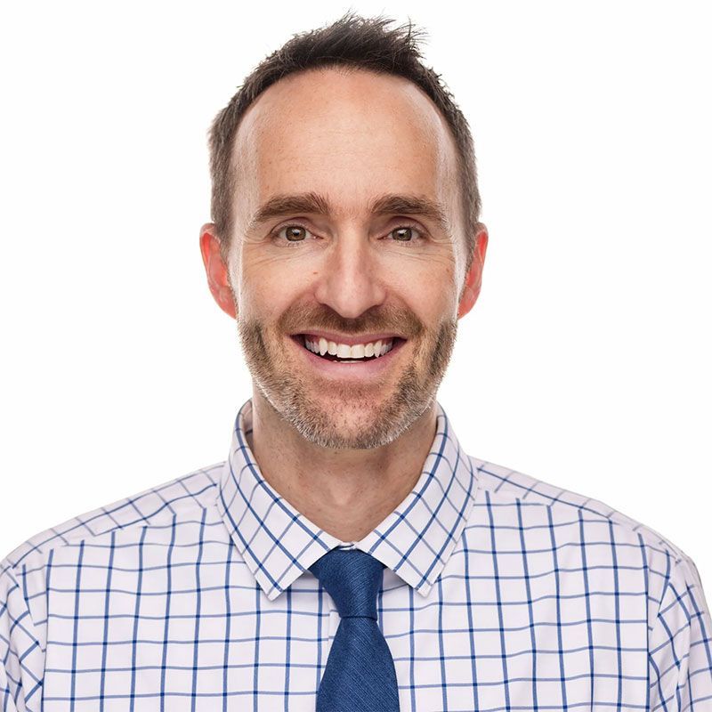 Smiling man with fair skin, wearing a blue tie, white and blue checkered shirt, against a white backdrop.