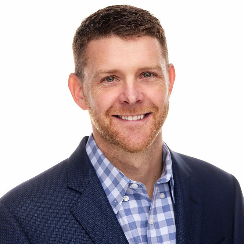 Smiling man with short brown hair, wearing a blue blazer and a light blue checkered shirt.