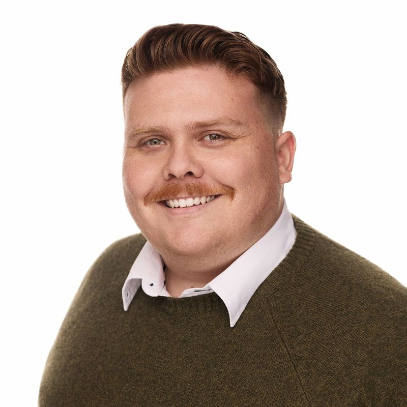 Man with a mustache smiles, wearing a green sweater over a white collared shirt. White background.