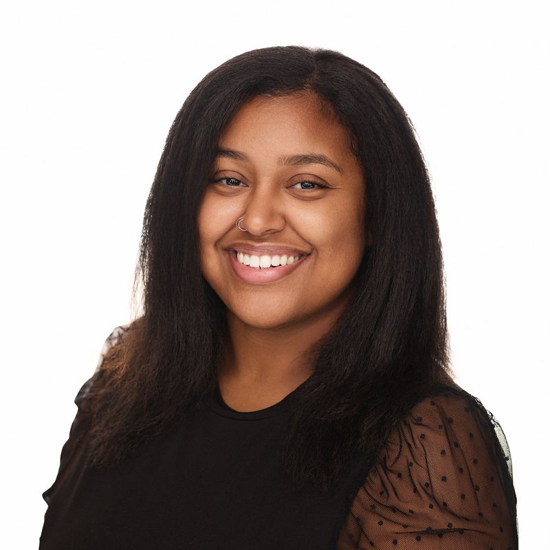 Woman with dark hair smiles, wearing a black top with sheer, polka-dot sleeves.