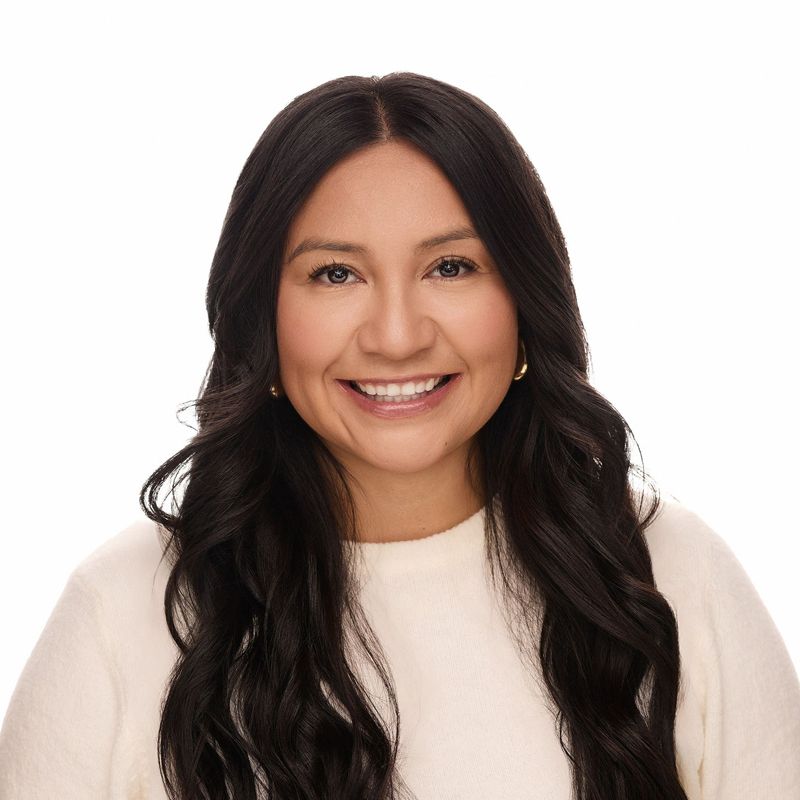 Woman with long dark hair smiles at the camera, wearing a cream-colored sweater. White background.