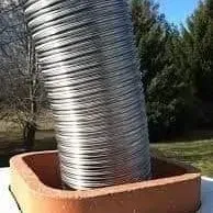 Flexible metal chimney liner extends from a terracotta chimney pot, outdoors in sunlight.